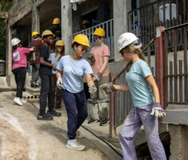 A group of people wearing hard hats and gloves work together carrying a cable outside a concrete building under construction.