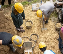 A group of people wearing yellow hard hats are shoveling and arranging gravel into piles, viewed from above.