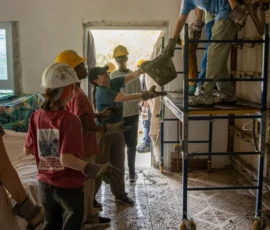 A group of people wearing safety gear work together indoors, passing debris in a line; one person stands on scaffolding handing a bag down.