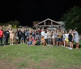 A large group of people, mostly young, pose together outdoors at night in front of a wooden shelter, standing on grass.