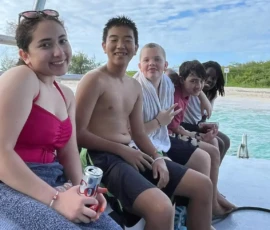 Five kids sit on the edge of a boat by the water, some smiling at the camera, wrapped in towels and swimwear, with trees and a partly cloudy sky in the background.