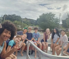 A group of kids and one adult sit and pose for a photo on an outdoor boat or open vehicle, with trees and cloudy sky in the background.
