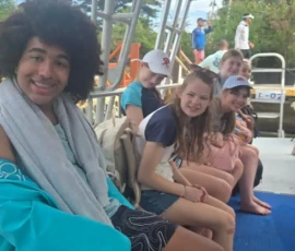 A group of young people sit on a boat, some smiling at the camera, with trees and more people visible in the background.