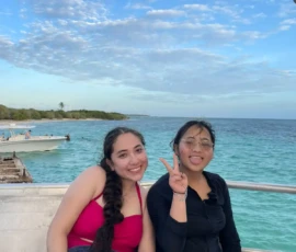 Two young women sit on a dock by the ocean, one wearing a pink swimsuit and towel, the other in black with a peace sign, with clear blue water and sky in the background.