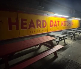 Outdoor view of picnic tables in front of a yellow and red sign reading "Heard Dat Kitchen New Orleans Best Kept Secret" at dusk.