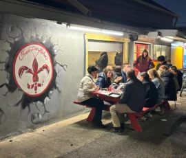 A group of people sit around a red picnic table eating outside a building at night, near a wall mural featuring a red fleur-de-lis inside a cracked circle.