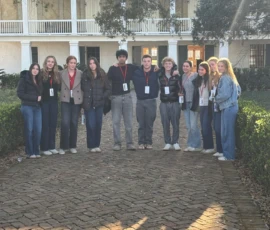 Twelve people wearing conference lanyards stand in a line on a brick path in front of a two-story house with columns, surrounded by greenery.