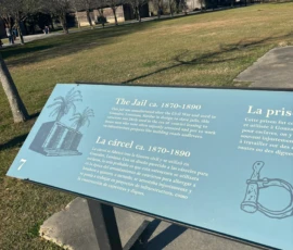 Outdoor informational sign titled "The Jail ca. 1870-1890" with text in English and Spanish, illustrations of a palm tree, a jail cell, and handcuffs, set in a park area.