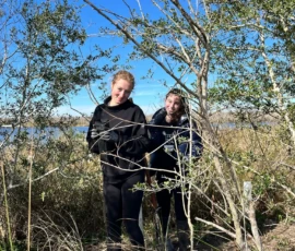 Two people stand among tall grass and shrubs near a body of water under a clear blue sky.