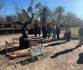 A group of people stand near a small winged statue on a brick patio under a clear blue sky, with trees and palm trees in the background.