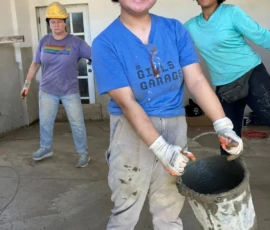 Three people wearing hard hats and work gloves stand indoors on a construction site; one smiles at the camera holding a bucket filled with wet concrete.