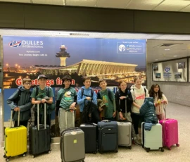 Seven children with luggage stand in front of a Dulles International Airport sign inside an airport terminal.