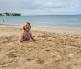 A child in a pink swimsuit lies partially buried in sand on a beach, smiling at the camera, with the ocean and cloudy sky in the background.