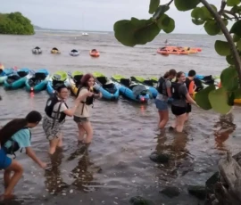 A group of people wearing life jackets stand and walk in shallow water near a shore with many colorful kayaks lined up on the sand.
