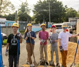 Five children wearing construction helmets and gloves stand on a street with shovels, next to buckets filled with gravel.