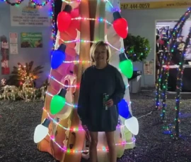 A person stands in front of a brightly lit, cone-shaped Christmas decoration with colorful lights and a large green star on top, at night.
