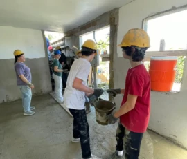A group of people wearing hard hats work together inside a building, passing buckets of material and smoothing concrete on the walls and floor.
