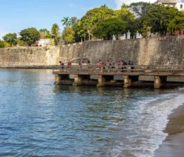 A wooden pier extends over the water near a stone wall, with several people sitting on the pier and trees visible in the background under a clear blue sky.