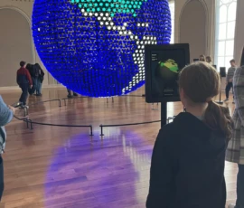 A girl stands in front of an interactive screen with a large digital globe display in a museum gallery; visitors are seen in the background.