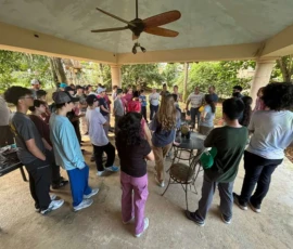 A group of people gathered under a covered outdoor patio, standing and talking near a table, surrounded by trees and greenery.