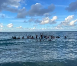 A group of people stand in the ocean with their arms raised, under a mostly blue sky with scattered clouds.