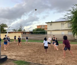 A group of children play a game of volleyball on a sandy outdoor court, with some players preparing to hit the ball over the net. Residential buildings are visible in the background.