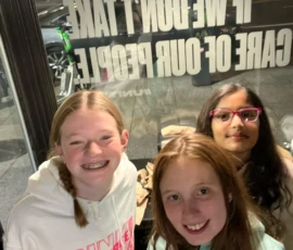 Three girls smile and pose indoors near a window with bold white text printed on the glass, while a street scene is visible outside at night.