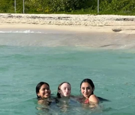 Three young people are swimming close together in clear, shallow ocean water near a sandy beach with greenery and two signs visible in the background.