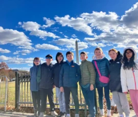 A group of eight children in winter clothing stand in front of a fence with the Washington Monument and blue sky in the background.