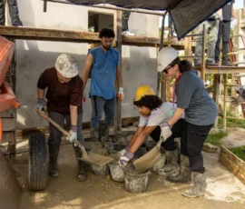 A group of people wearing hard hats and gloves work together to shovel and collect wet cement into buckets at a construction site.