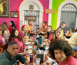 A group of young people sits around a table enjoying a meal in a colorful, busy restaurant with vibrant pink and green walls.