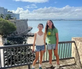 Two young women stand on a stone lookout by the water, with a historic fort and a blue sky with clouds in the background.
