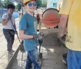 A child wearing a hard hat, gloves, and boots stands on wet cement, holding a tool, while other people work around him at a construction site.