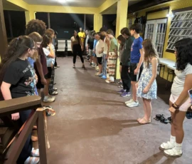 A group of people stands in two facing lines on a covered patio, looking down at the floor, with a person in black standing at the center.