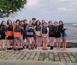 A group of people wearing life jackets stand together on a paved area near the shoreline, with the ocean and a boat in the background.