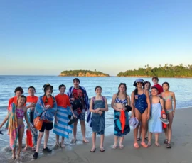 A group of teenagers and children stand on a sandy beach near the water, some wrapped in towels, with an island and clear sky in the background.