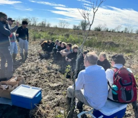 A group of people sit and stand in a grassy, open field under a blue sky, some with backpacks and coolers, appearing to listen to a speaker.