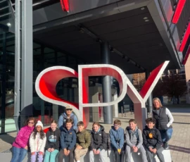 A group of children and two adults pose in front of a large red and white "SPY" sign outside a modern building.