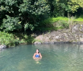 A person is standing in a natural pool surrounded by green trees and rocks on a sunny day.