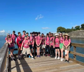 A group of teenagers in matching pink shirts stand on a wooden boardwalk by the water, posing for a photo under a clear blue sky.