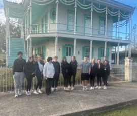 A group of twelve people stands on the sidewalk in front of a light blue, two-story house with ornate decorations and balconies.