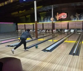 A person bowls a green ball down a lane at an indoor bowling alley with neon signs and retro decor.