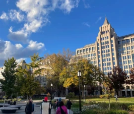 People walk along a paved path in an urban park, with tall office buildings and autumn trees under a blue sky with scattered clouds.
