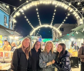 Four women stand together at an outdoor night market under arching string lights, surrounded by vendor booths and illuminated artwork displays.