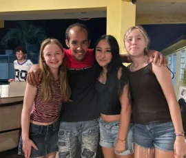Four people stand close together, smiling for a photo in an outdoor, covered area at night. Another person is visible in the background near a counter.