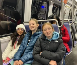 Three girls in winter jackets sit together on a subway train, with other passengers seated behind them. The train interior is visible.