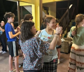A group of teenagers practice partner dancing indoors, with two girls in the foreground holding hands and following dance steps. Other pairs are visible in the background.