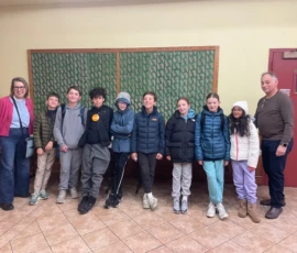A group of eight children and two adults stand indoors against a wall with a green bulletin board, all dressed in casual winter clothing.