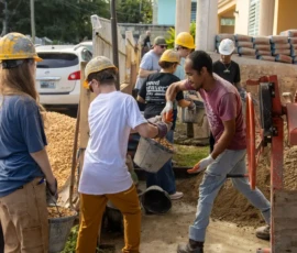 A group of people wearing hard hats work together, passing buckets of dirt and gravel at a construction site outdoors.