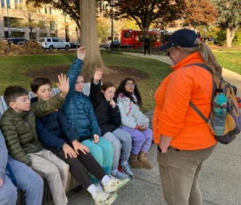 A group of children sit on a bench outdoors, some raising their hands, while an adult in an orange jacket stands facing them.